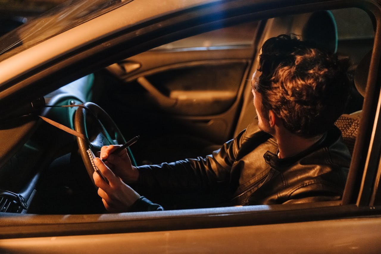 services-03 A man in a leather jacket taking notes inside a car at night, implying a stakeout or investigation setting.