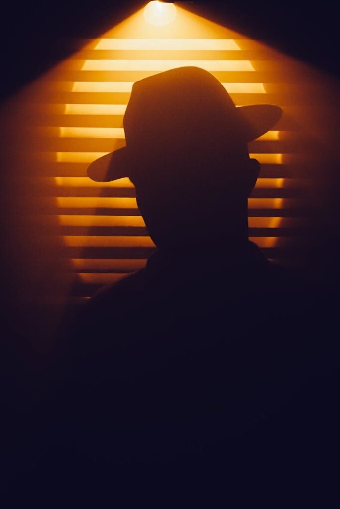 pexels photo 15326392 Mysterious silhouette of person in hat against a backdrop of dramatic blinds and warm lighting.