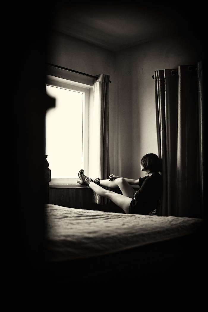 services-02 A young woman sits pensively by a window in a serene black and white bedroom.