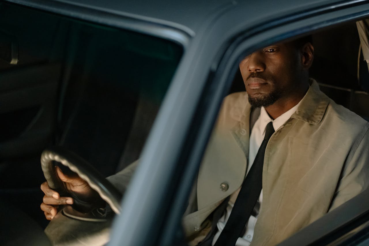our-experience A man in a trench coat sits inside a car, reflecting a serious demeanor through the window.